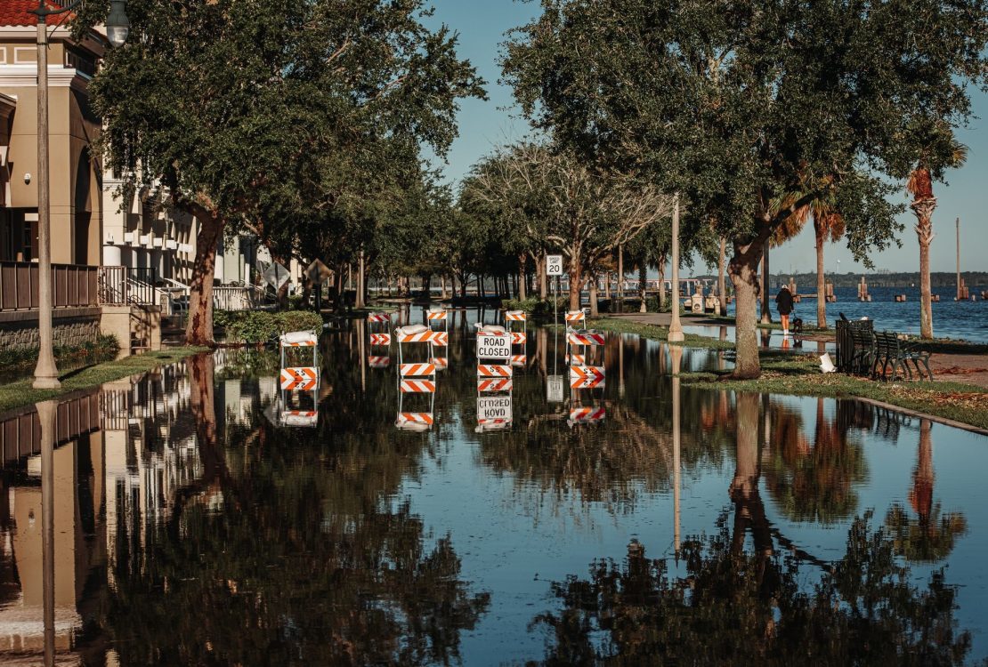 Rua alagada com placas de interdição refletidas na água ao lado de uma calçada arborizada.
