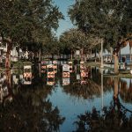 Rua alagada com placas de interdição refletidas na água ao lado de uma calçada arborizada.