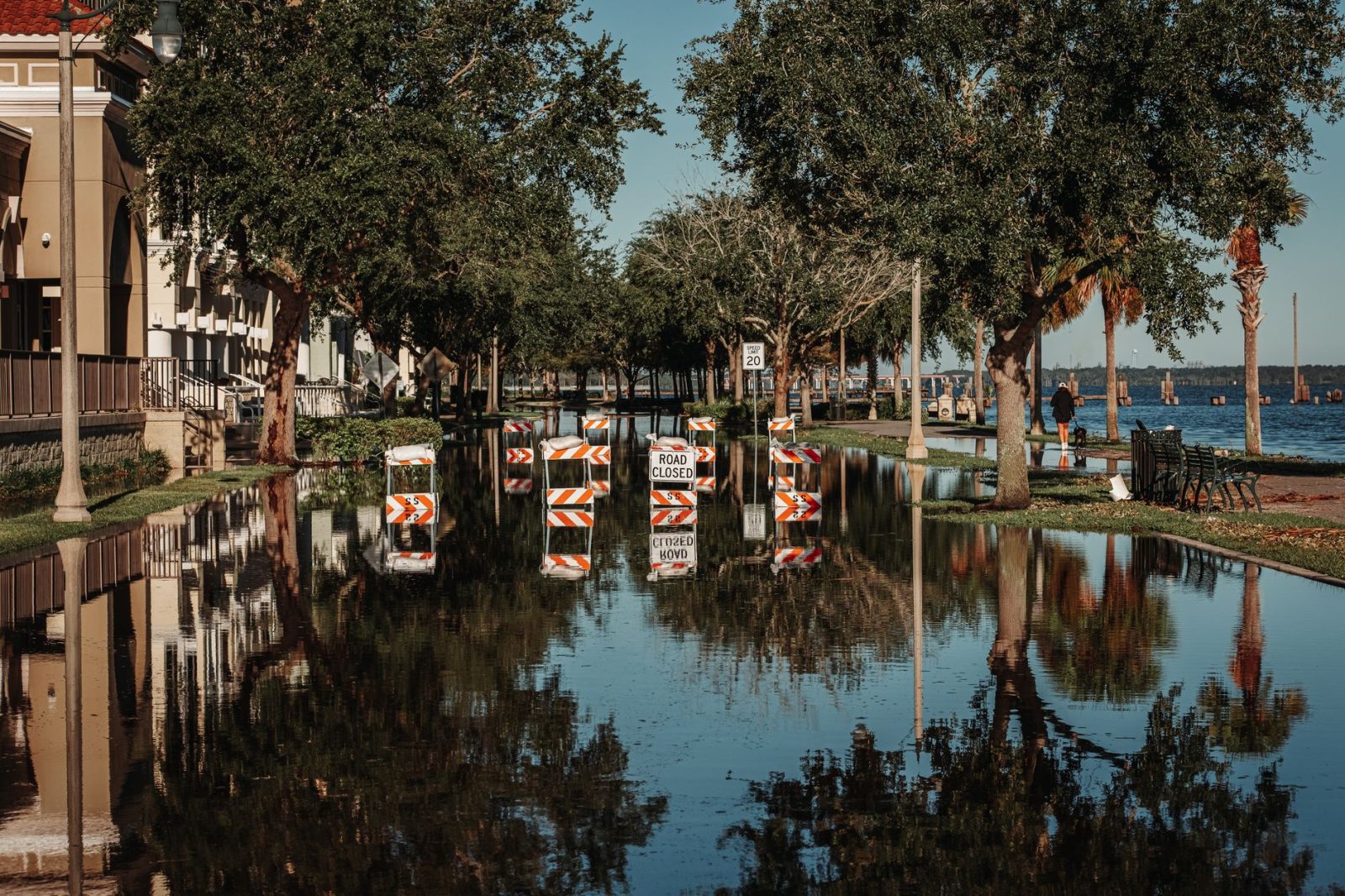Rua alagada com placas de interdição refletidas na água ao lado de uma calçada arborizada.