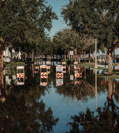 Rua alagada com placas de interdição refletidas na água ao lado de uma calçada arborizada.